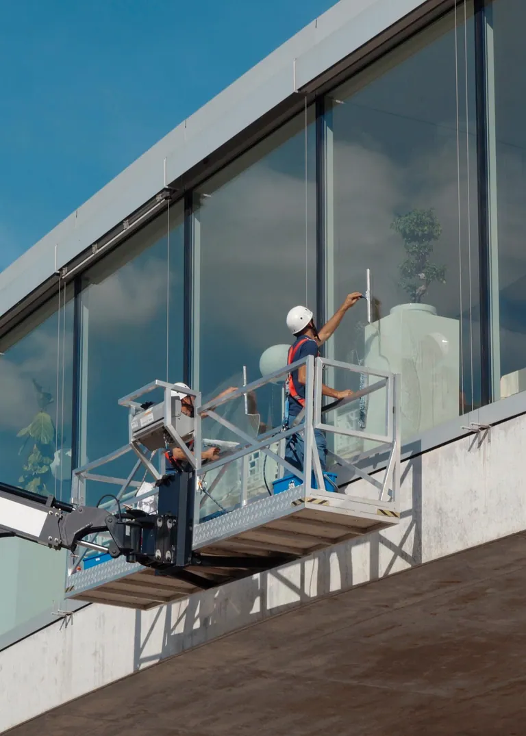 Zwei Reinigungskräfte reinigen große Fassadenfenster vom Korb einer Hebebühne aus.